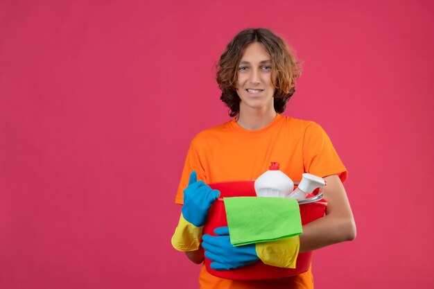 Jovem com camiseta laranja usando luvas de borracha, segurando um balde com ferramentas de limpeza, olhando para a câmera, sorrindo, positivo e feliz em pé sobre o fundo rosa