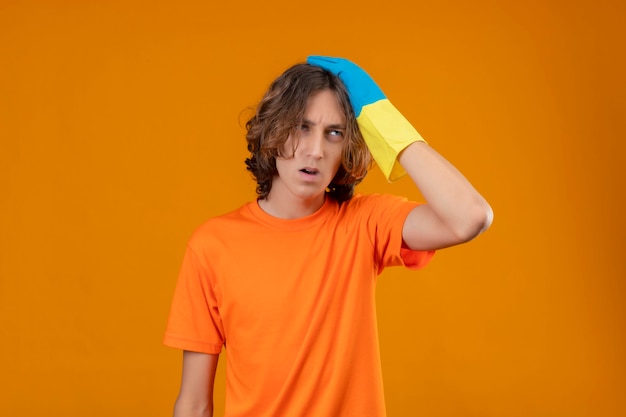 Jovem com camiseta laranja usando luvas de borracha tocando a cabeça por engano, parecendo confuso com conceito de memória ruim em pé sobre fundo amarelo