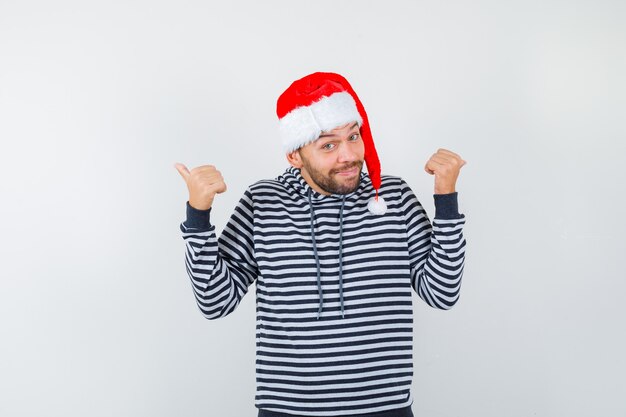 Jovem com capuz, chapéu de Papai Noel apontando os polegares para trás e parecendo alegre,