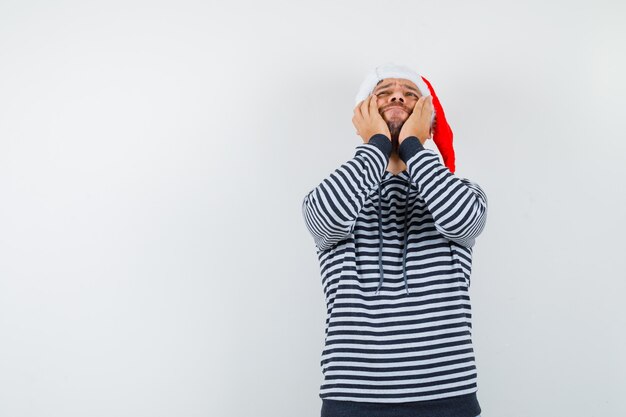 Jovem com capuz, chapéu de papai noel, mantendo as mãos nas bochechas, olhando para cima e parecendo esperançoso,