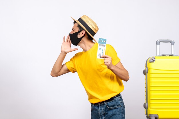 Jovem com chapéu de palha em frente a uma mala amarela segurando uma passagem chamando algo