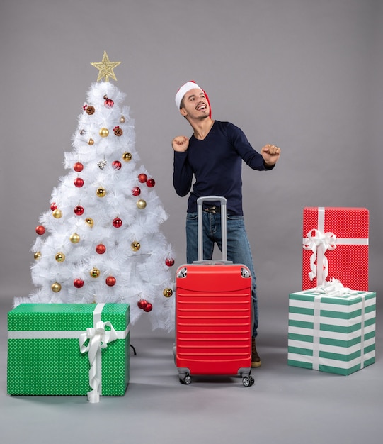 Jovem com chapéu de papai noel em pé perto da árvore de natal branca e presentes coloridos em cinza.