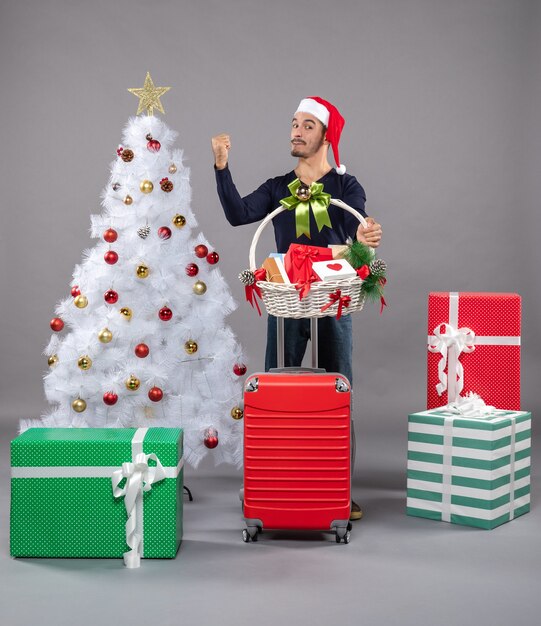 Jovem com chapéu de Papai Noel segurando uma cesta de presentes e mostrando a árvore de natal em cinza isolado