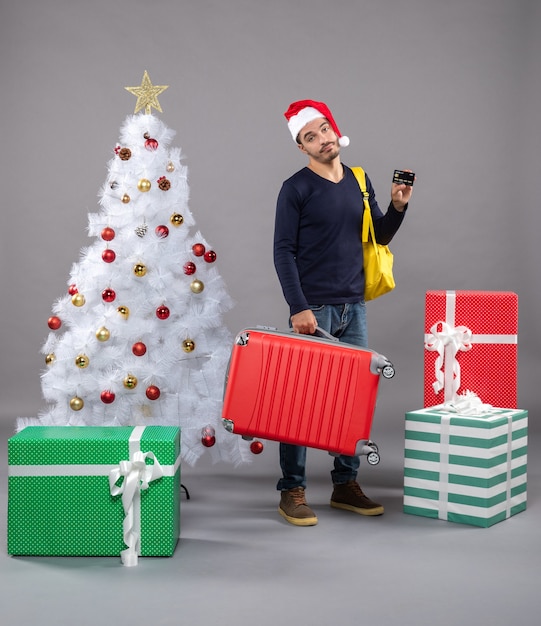 Jovem com chapéu de papai noel segurando uma mala vermelha e mostrando um cartão em torno de presentes isolados.