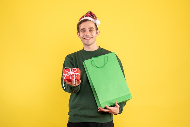 Jovem com chapéu de Papai Noel segurando uma sacola de compras verde e um presente em pé no espaço de cópia com fundo amarelo