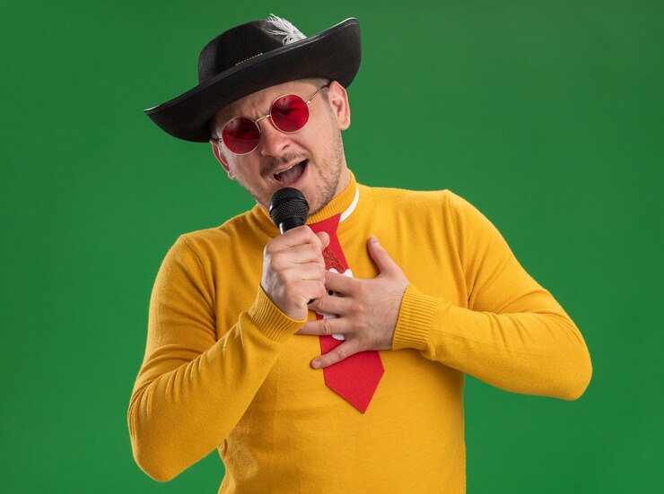Foto grátis jovem com gola amarela e óculos com gravata vermelha engraçada cantando segurando o microfone feliz e alegre em pé sobre a parede verde
