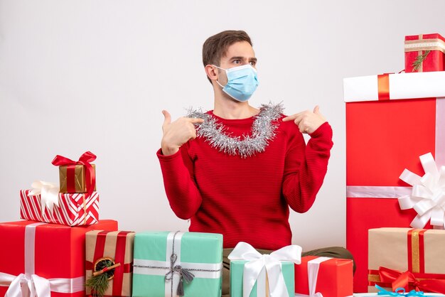 Jovem com máscara apontando para as costas sentado em volta dos presentes de natal