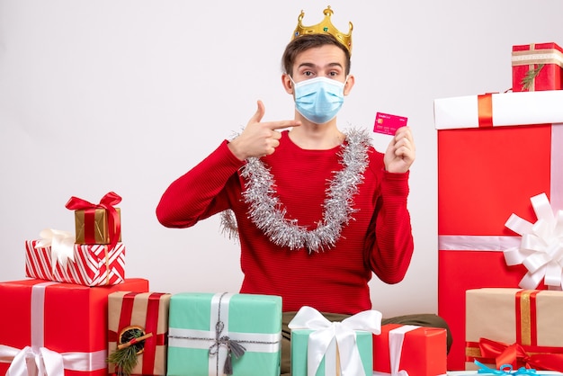 Jovem com máscara apontando para um cartão sentado no chão. Presentes de Natal