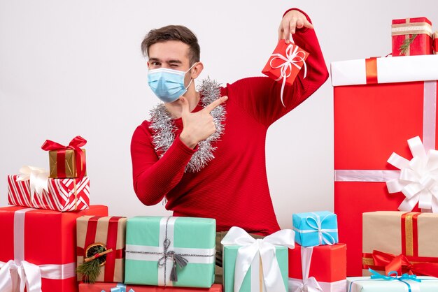 Jovem com máscara apontando para um presente próximo aos presentes de Natal