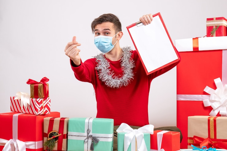 Foto grátis jovem com máscara fazendo gesto de coração com dedo, sentado no chão, presentes de natal