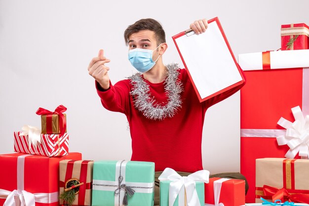 Jovem com máscara fazendo gesto de coração com dedo, sentado no chão, presentes de natal