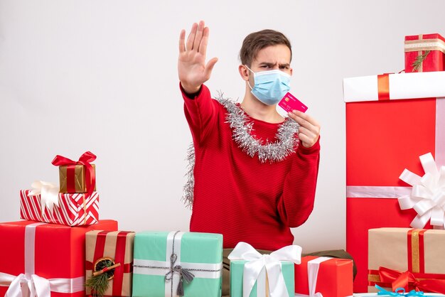 Jovem com máscara fazendo sinal de pare sentado em frente aos presentes de natal