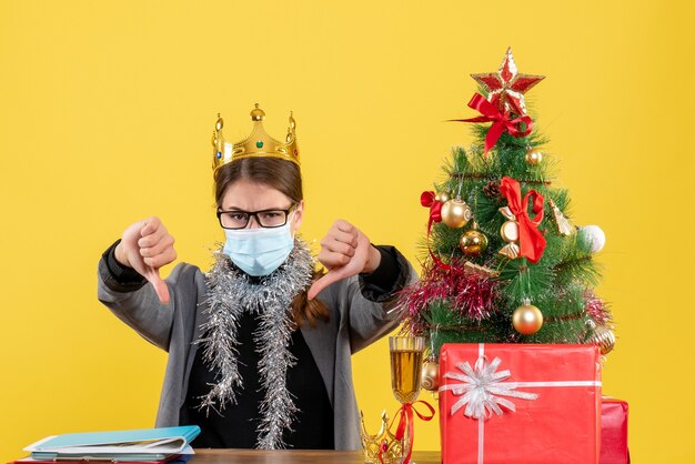 Jovem com máscara médica fazendo gesto com o polegar para baixo, árvore de natal e presentes