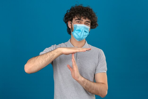 Jovem com máscara protetora médica parecendo doente e cansado fazendo gesto de tempo com as mãos sobre fundo azul isolado