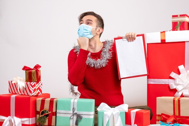 Jovem com máscara segurando uma prancheta sentado em frente a uma foto de presentes de natal