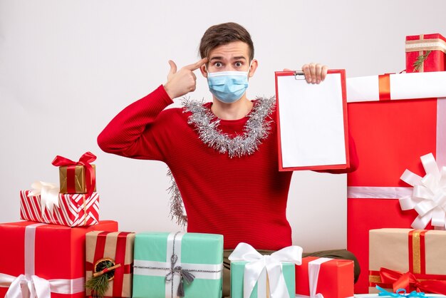 Jovem com máscara segurando uma prancheta sentado em frente aos presentes de natal