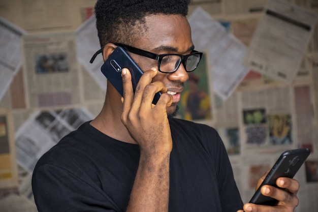 Jovem com óculos falando ao telefone enquanto usa outro em uma sala