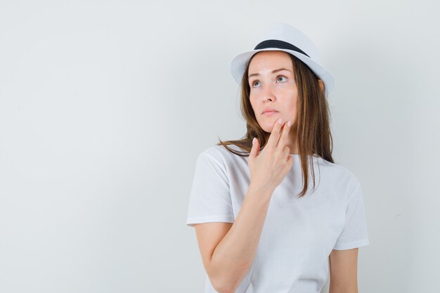 Jovem com os dedos no queixo com um chapéu de camiseta branca e parecendo pensativa