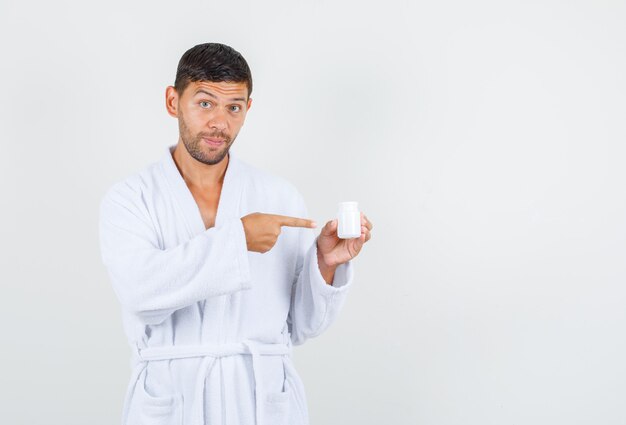 Jovem com roupão branco apontando para o frasco de remédio de plástico, vista frontal.