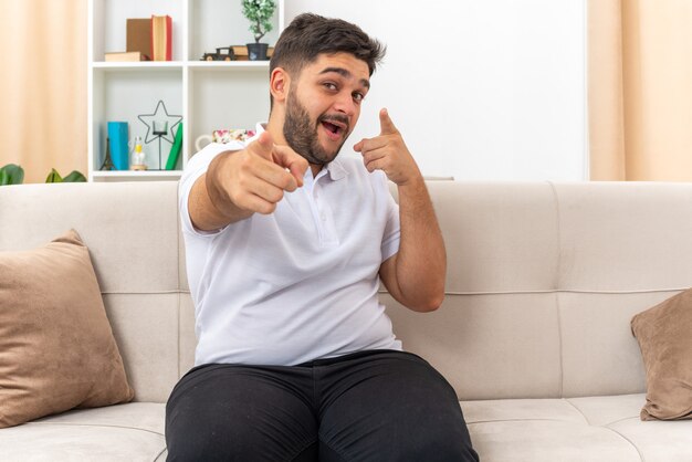 Jovem com roupas casuais apontando com o dedo indicador para a frente, sorrindo, confiante, feliz e positivo, sentado em um sofá na sala de estar iluminada