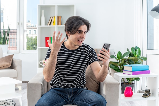 Jovem com roupas casuais com smartphone parecendo espantado e surpreso sentado na cadeira na luz da sala de estar