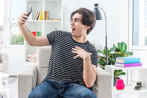 Jovem com roupas casuais fazendo selfie usando o smartphone parecendo espantado e surpreso sentado na cadeira na sala iluminada