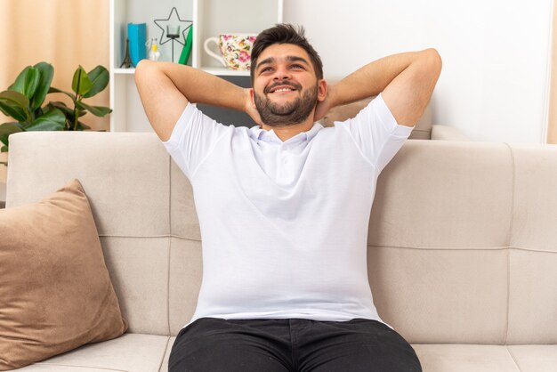 Jovem com roupas casuais feliz e positivo sorrindo alegremente relaxando sentado em um sofá na sala de estar iluminada