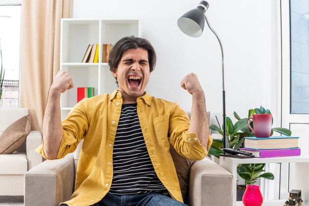 Jovem com roupas casuais, louco, feliz e animado, gritando com os punhos cerrados, sentado na cadeira na sala iluminada