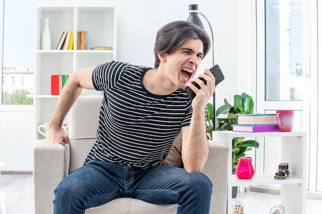 Jovem com roupas casuais, parecendo animado e frustrado, gritando ao falar no celular, sentado na cadeira na sala iluminada