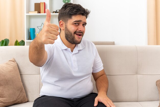 Jovem com roupas casuais, parecendo feliz e alegre, mostrando os polegares para cima, sentado em um sofá na sala de estar iluminada