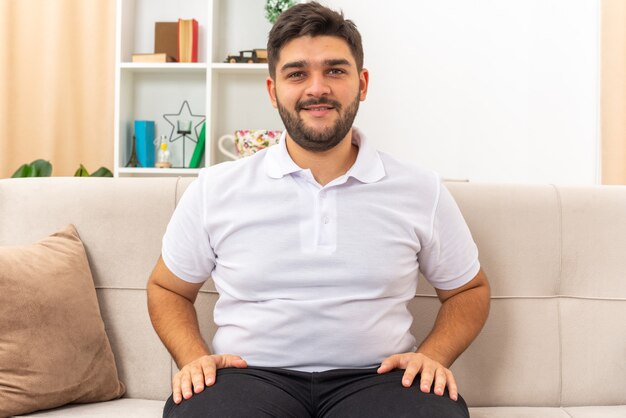 Jovem com roupas casuais, parecendo feliz e otimista, passando o fim de semana em casa sentado em um sofá na sala de estar iluminada