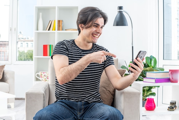 Jovem com roupas casuais segurando um smartphone, olhando para ele, feliz e alegre, sentado na cadeira na luz da sala de estar