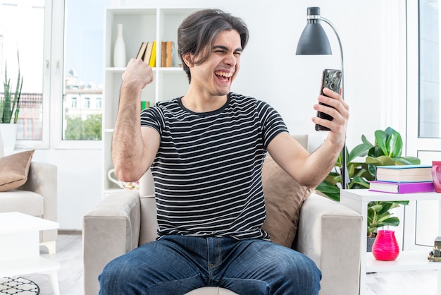 Jovem com roupas casuais segurando um smartphone, olhando para ele feliz e animado, levantando o punho sentado na cadeira na sala iluminada