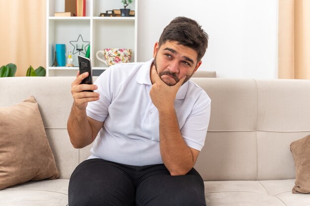 Jovem com roupas casuais segurando um smartphone, olhando para o lado, perplexo, sentado em um sofá em uma sala iluminada