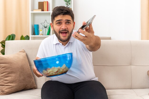 Jovem com roupas casuais segurando uma tigela de batatas fritas e um smartphone parecendo confuso sentado em um sofá na sala de estar iluminada