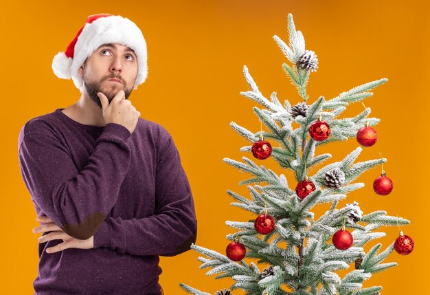 Jovem com suéter roxo e chapéu de Papai Noel olhando para cima com uma expressão pensativa no rosto pensando ao lado da árvore de Natal sobre fundo laranja