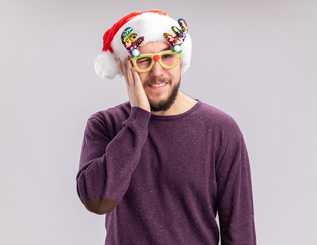 Jovem com suéter roxo e chapéu de Papai Noel usando óculos engraçados, olhando para a câmera feliz e positivo, sorrindo e piscando em pé sobre um fundo branco
