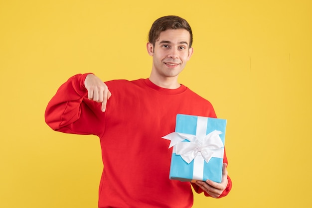 Jovem com suéter vermelho segurando uma caixa de presente azul em fundo amarelo de frente