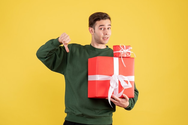 Jovem com um presente de natal fazendo uma placa com o polegar para baixo em pé sobre fundo amarelo.