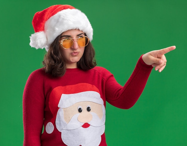 Jovem com um suéter de natal, usando óculos e chapéu de Papai Noel, olhando para o lado descontente, apontando com o dedo indicador para algo em pé sobre a parede verde