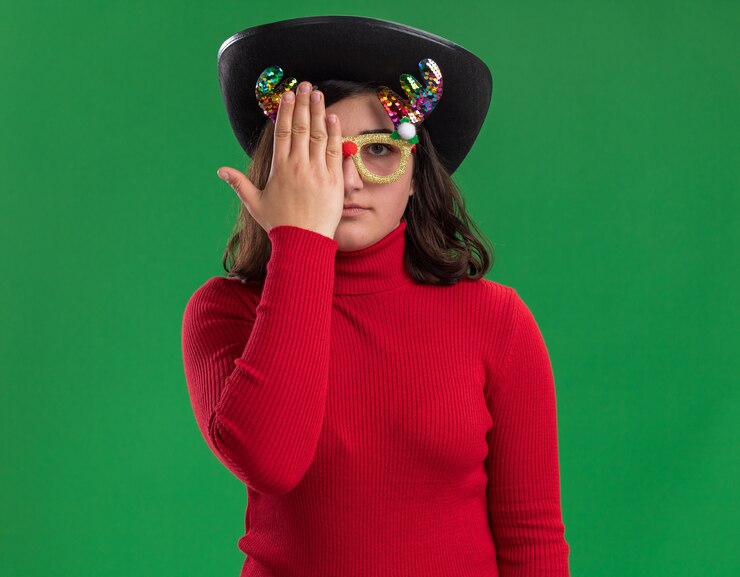 Foto grátis jovem com um suéter vermelho usando óculos engraçados e chapéu preto cobrindo um olho com a mão