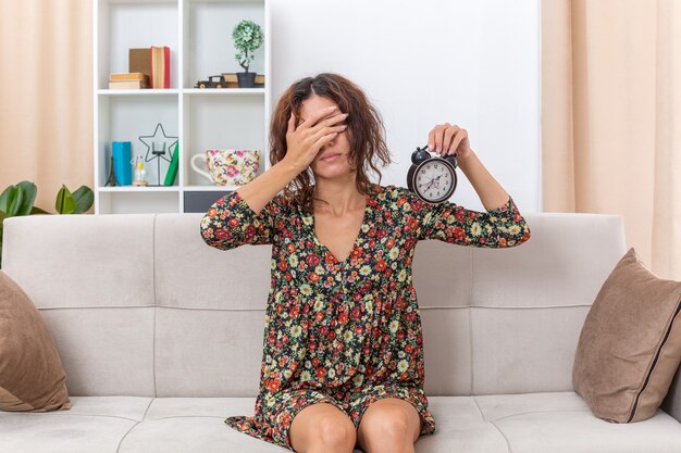 Jovem com um vestido floral segurando um despertador cobrindo os olhos com a mão cansada e entediada, sentada em um sofá na sala iluminada