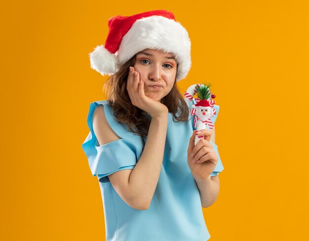 Jovem com uma blusa azul e um chapéu de Papai Noel segurando uma bengala de doces de Natal, parecendo confusa e descontente com a mão no queixo