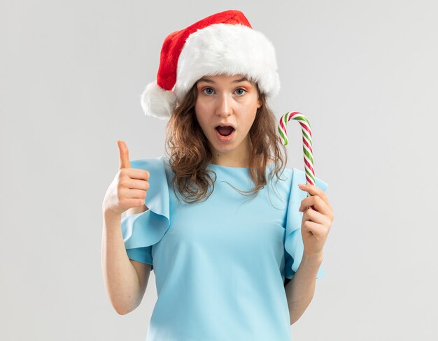 Jovem com uma blusa azul e um chapéu de Papai Noel segurando uma bengala de doces, parecendo feliz e surpresa, mostrando os polegares para cima