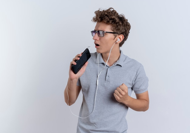 Foto grátis jovem com uma camisa pólo cinza com fones de ouvido segurando um smartphone usando como microfone cantando em pé sobre uma parede branca