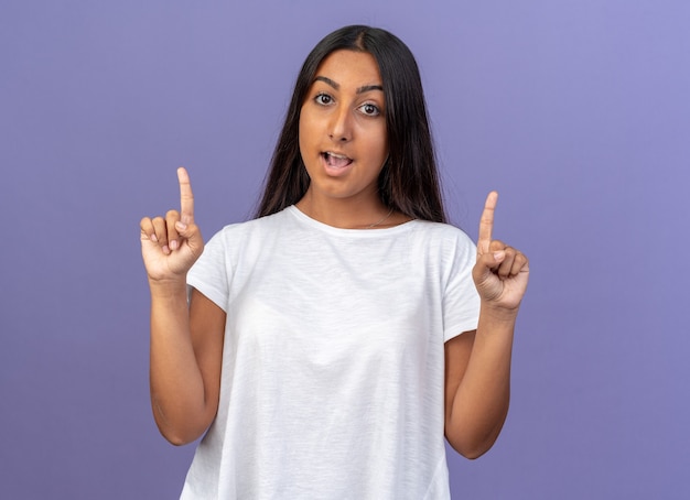 Jovem com uma camiseta branca olhando para a câmera feliz e surpresa, mostrando os dedos indicadores, tendo uma nova ideia