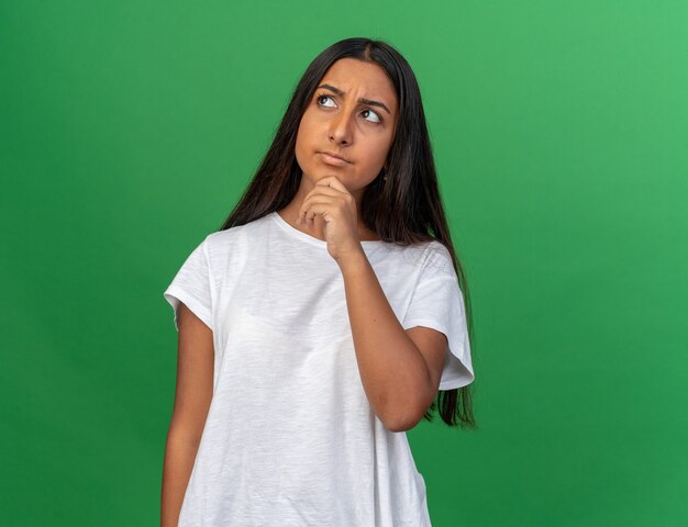 Jovem com uma camiseta branca olhando para cima intrigada com a mão no queixo parada sobre um fundo verde