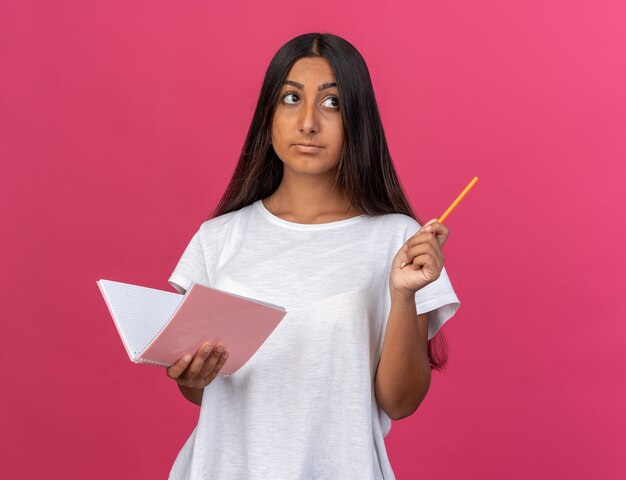 Jovem com uma camiseta branca segurando um caderno e um lápis olhando para o lado intrigada