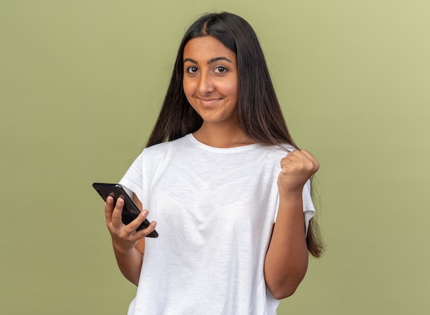 Jovem com uma camiseta branca segurando um smartphone, olhando para a câmera, feliz e animada, cerrando os punhos