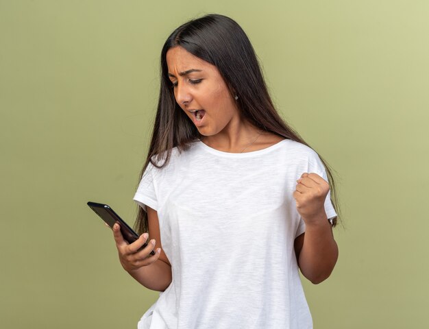 Jovem com uma camiseta branca segurando um smartphone olhando para ele com expressão agressiva cerrando o punho em pé sobre um fundo verde